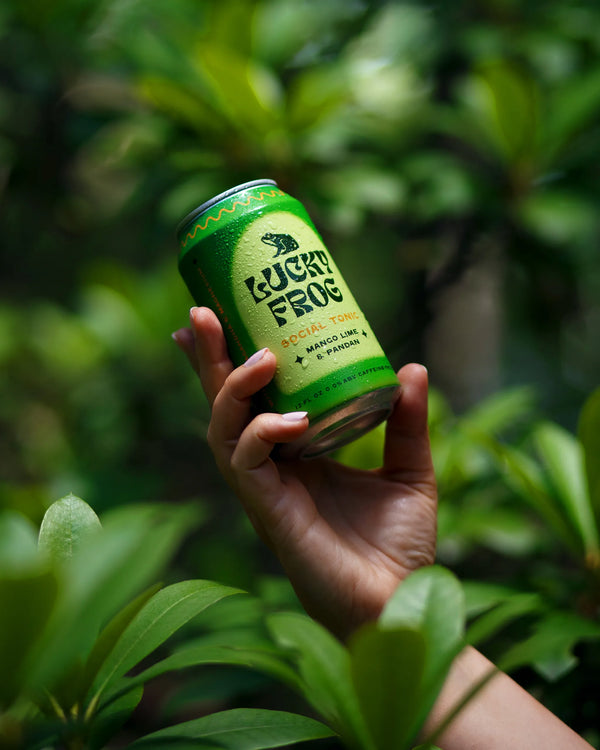 Hand holding a can of Lucky Frog Mango Lime & Pandan social tonic amid green leafy plants