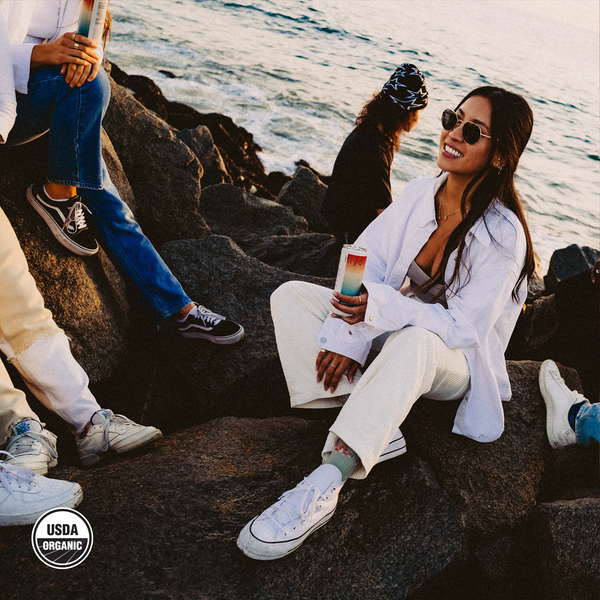 hiyo Watermelon Lime alcohol-free seltzer can held by a woman sitting on coastal rocks by the ocean
