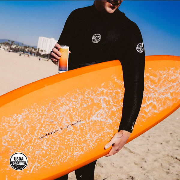 Surfer in wetsuit holding a hiyo Peach Mango alcohol-free functional seltzer can with orange surfboard on beach