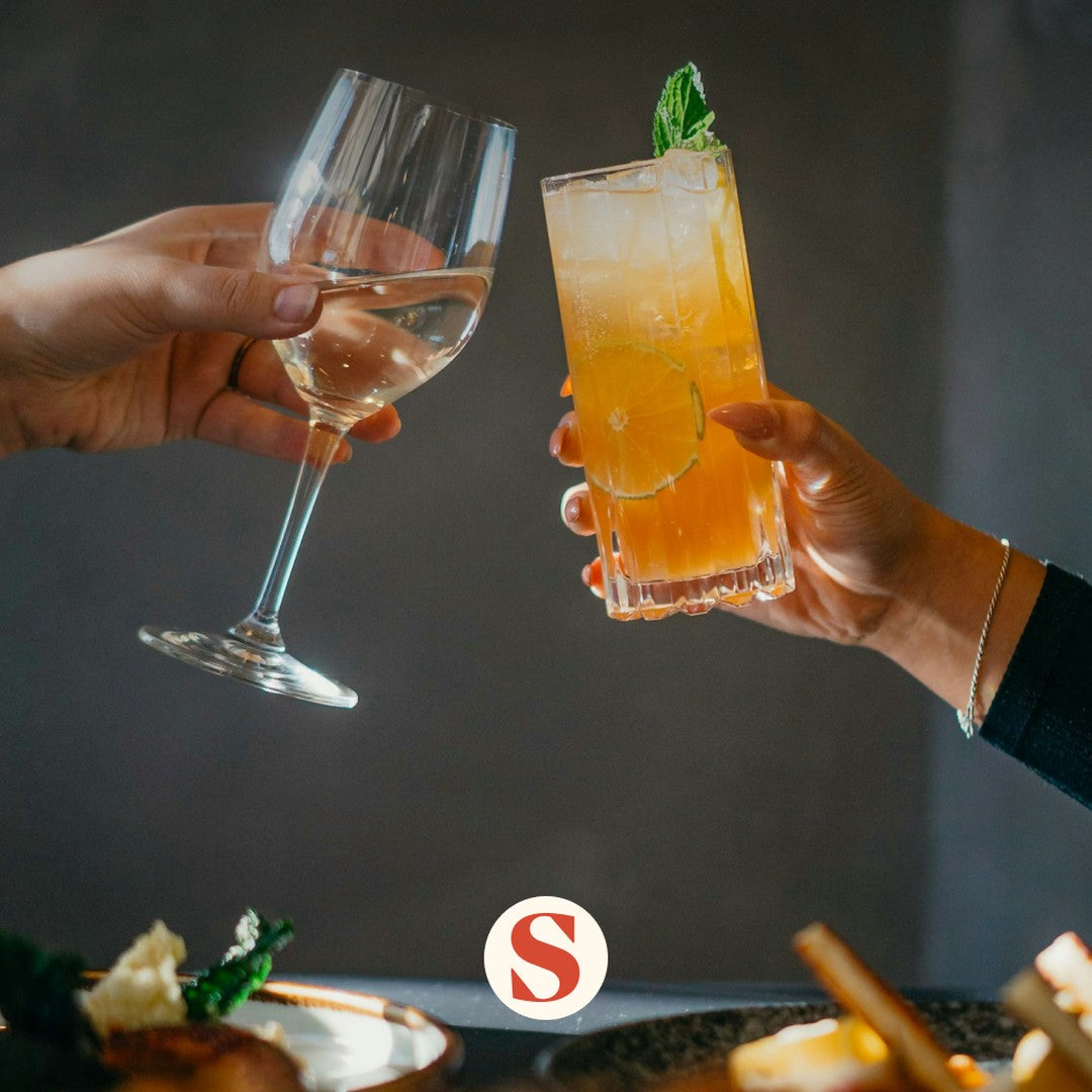 Two people clinking a white wine glass and an orange alcohol-free cocktail at Swizzle
