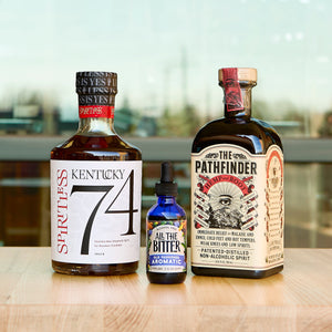 Three bottles on a wooden table with a blurred indoor background. Kentucky 74 AF bourbon, All the Bitter Old Fashioned Aromatic, The Pathfinder