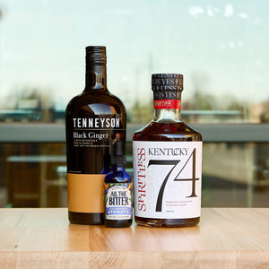 A lifestyle photograph featuring the Ginger Old Fashioned Cocktail Kit bottles on a light wood surface. The lineup includes a tall, dark bottle of Tenneyson Black Ginger, a square bottle of Spiritless Kentucky 74 non-alcoholic bourbon, and a small blue glass dropper bottle of All The Bitter Old Fashioned Aromatic bitters. The background is a bright, softly blurred indoor setting with large windows.