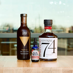 A lifestyle photograph featuring the Maple Old Fashioned Cocktail Kit bottles on a light wood surface. The lineup includes a matte brown bottle of Three Spirit Nightcap, a small blue glass dropper bottle of All The Bitter Spiced Cherry bitters, and a bottle of Spiritless Kentucky 74 non-alcoholic bourbon. The background is a bright, softly blurred indoor setting with large windows.