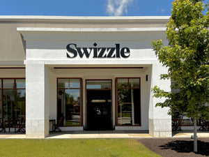 Exterior view of Swizzle Market storefront featuring open double doors, large glass windows, modern signage, and a leafy tree in the foreground.