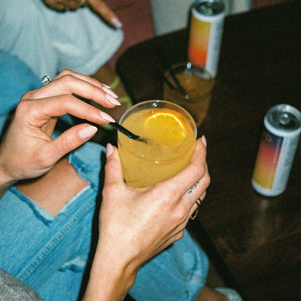 Hands holding a glass of hiyo Peach Mango alcohol-free functional seltzer with orange slice, cans nearby