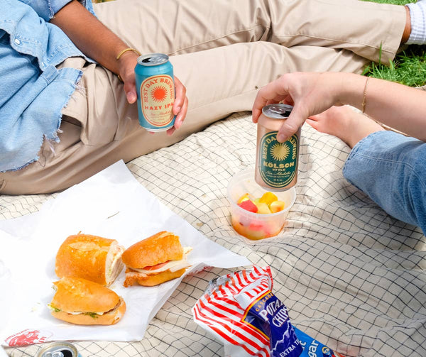 Two Best Day non-alcoholic beer cans held at a picnic with sandwiches on a blanket